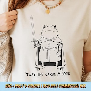 May include: Beige t-shirt featuring a black and white illustration of a frog dressed as a knight, holding a sword. The text "'TWAS THE CARBS M'LORD" is printed below the frog. The shirt is a casual wear item.