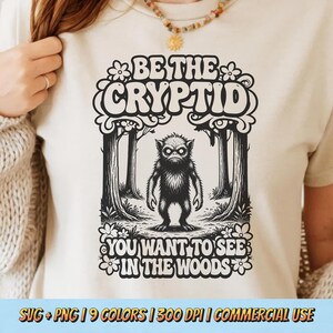 Pode incluir: Camiseta bege com um gráfico em preto e branco de uma criatura críptica em um cenário de floresta. O texto diz "BE THE CRYPTID YOU WANT TO SEE IN THE WOODS". O design inclui elementos florais.