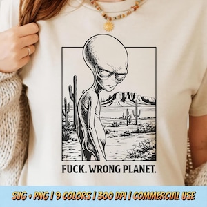 Puede incluir: Camiseta beige con una ilustración en blanco y negro de un extraterrestre con expresión enfadada, en un paisaje desértico. El texto "FUCK. WRONG PLANET." está debajo de la imagen.