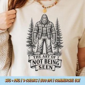 May include: A cream-colored t-shirt featuring a black graphic of a Bigfoot creature wearing a plaid shirt and jeans, standing between two pine trees. Below the creature is a banner with the text THE ART OF NOT BEING SEEN in black lettering.