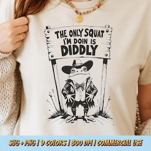 May include: A cream-colored t-shirt featuring a black and white illustration of a frog dressed in a suit and cowboy hat. The frog stands in front of a sign that reads "THE ONLY SQUAT I'M DOIN IS DIDDLY".