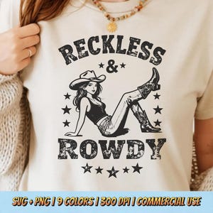 Op de afbeelding: Crèmekleurig T-shirt met een zwart-witte afbeelding. De afbeelding toont een vrouw met een cowboyhoed en laarzen, met de woorden "RECKLESS & ROWDY" erboven en eronder. Sterren omringen de afbeelding.