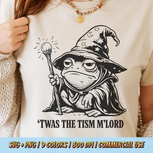 May include: A cream-colored t-shirt featuring a black and white illustration of a frog dressed as a wizard, holding a staff. The frog wears a pointed hat with stars and a crescent moon. The text "'TWAS THE TISM M'LORD" is printed below the illustration.