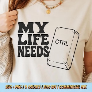 Può includere: T-shirt beige con la scritta "MY LIFE NEEDS" in grassetto nero. Una grande grafica in bianco e nero di un tasto "CTRL" è accanto al testo. La maglietta è di colore neutro e ha maniche corte.