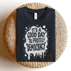 May include: Black t-shirt with the text "It's a good day to protect democracy" in white, within a white splatter design. The shirt is displayed on a woven, circular surface.