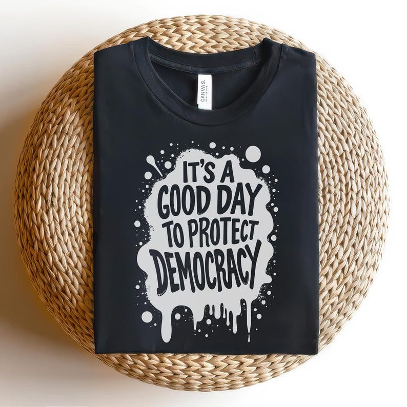 May include: Black t-shirt with the text "It's a good day to protect democracy" in white, within a white splatter design. The shirt is displayed on a woven, circular surface.