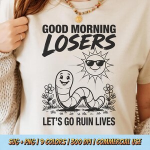 Pode incluir: Uma t-shirt de cor creme com uma impressão gráfica preta. O texto "GOOD MORNING LOSERS" aparece acima de um verme sorridente e um sol com óculos de sol. Abaixo do gráfico, o texto "LET'S GO RUIN LIVES" inclui pequenos detalhes florais.