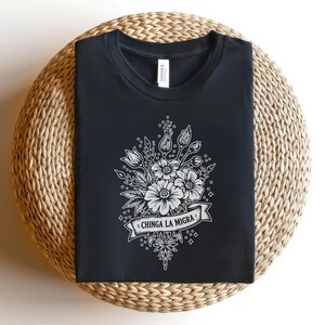 May include: Black t-shirt with a white floral design and the text "CHINGA LA MIGRA" on a banner. The design features detailed flowers, leaves, and buds. The shirt is displayed on a woven, circular surface.