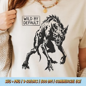May include: A cream-coloured t-shirt with a black illustration of a snarling, monstrous creature. The text "WILD BY DEFAULT" is in a black rectangular box. The shirt has short sleeves.