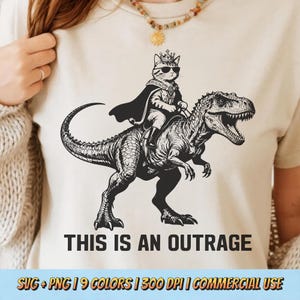 May include: Beige t-shirt featuring a black and white graphic of a cat wearing a crown and sunglasses, riding a Tyrannosaurus Rex. The text "THIS IS AN OUTRAGE" is below the graphic.