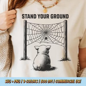 Könnte beinhalten: Beiges T-Shirt mit einer schwarz-weißen Grafik eines Schweins, das einem Spinnennetz mit einer Spinne gegenübersteht. Der Text "STAND YOUR GROUND" steht über der Grafik. Der untere Teil des Shirts trägt den Text "SVG + PNG | 9 COLORS | 300 DPI | COMMERCIAL USE".