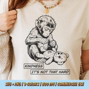 Puede incluir: Camiseta beige con una ilustración en blanco y negro de un mono cosiendo un oso de peluche. El texto "KINDNESS IT'S NOT THAT HARD" está debajo. La camiseta es de color neutro.