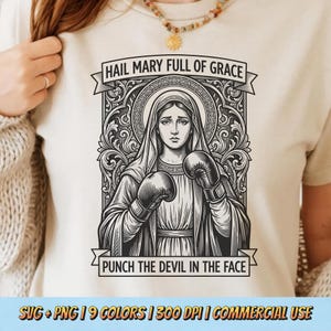 May include: Cream-colored t-shirt featuring a black and white illustration of a woman wearing boxing gloves. The text "HAIL MARY FULL OF GRACE" is above the image, and "PUNCH THE DEVIL IN THE FACE" is below. The shirt also includes the text "SVG + PNG | 9 COLORS | 300 DPI | COMMERCIAL USE".