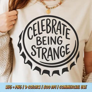 Peut inclure: T-shirt beige avec un motif noir circulaire représentant un bouchon de bouteille et l'inscription "CELEBRATE BEING STRANGE". La personne qui porte le t-shirt a les cheveux bruns et porte un collier.