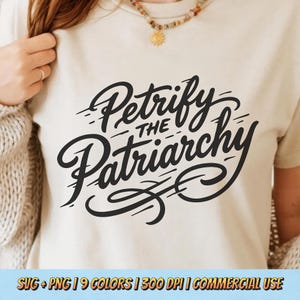 May include: A cream-colored t-shirt with the phrase "Petrify the Patriarchy" in black script. The text is in a stylized, cursive font. The shirt is being worn by a person, with a necklace visible at the neckline.