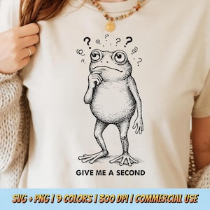 Puede incluir: Una camiseta color crema con una ilustración en blanco y negro de una rana en pose pensativa, con signos de interrogación sobre su cabeza. El texto "GIVE ME A SECOND" está impreso debajo. La camiseta es parte de una colección con múltiples opciones de color.