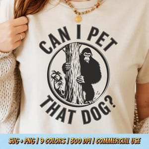 以下が含まれることがあります： 木の後ろから覗くゴリラの黒いグラフィックが描かれたベージュ色のTシャツ。周りには「CAN I PET THAT DOG？」の文字。ヤシの木のデザインも含まれており、ペットの飼育をユーモラスに表現しています。