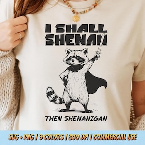 Può includere: T-shirt color crema con un'illustrazione in bianco e nero di un procione con mantello e spada. Il testo "I SHALL SHENANIGAN THEN SHENANIGAN" è stampato sopra e sotto il procione.