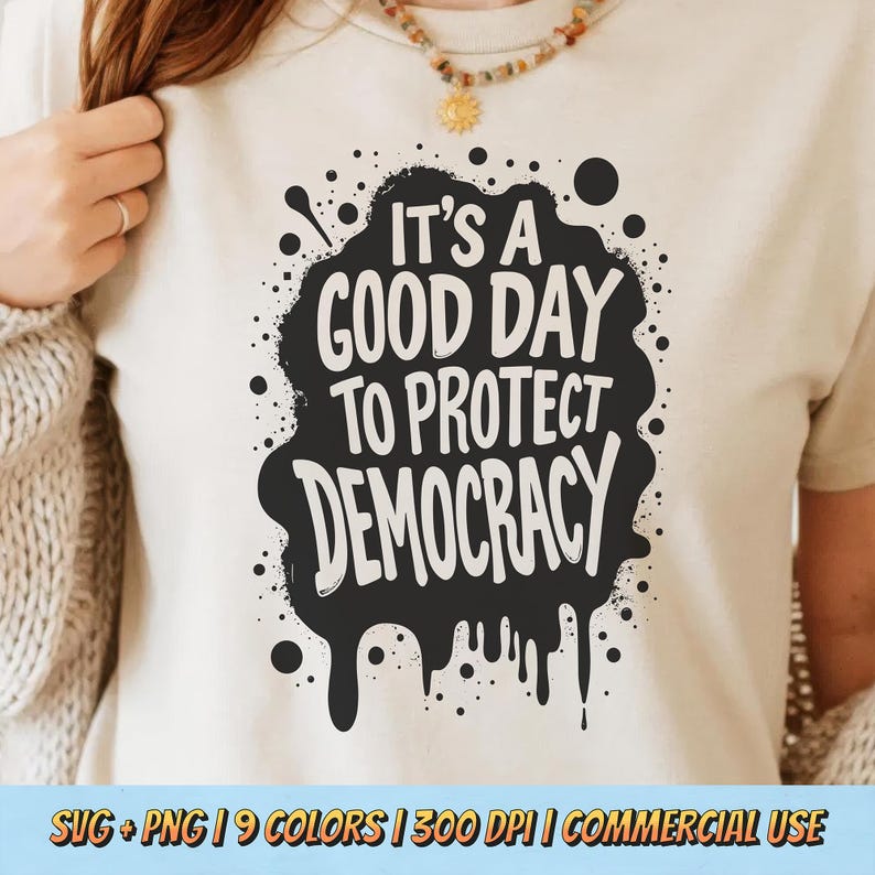 May include: A cream-colored t-shirt with the phrase "IT'S A GOOD DAY TO PROTECT DEMOCRACY" in bold white letters within a black, splattered design. The shirt also has additional text at the bottom.