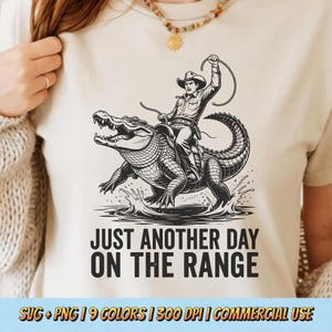 Op de afbeelding: Een beige t-shirt met een zwart-wit illustratie van een cowboy die op een alligator rijdt, met de tekst "JUST ANOTHER DAY ON THE RANGE". Het ontwerp bevat waterspetters en een touw. De onderkant van het shirt heeft de tekst "SVG + PNG | 9 COLORS | 300 DPI | COMMERCIAL USE."