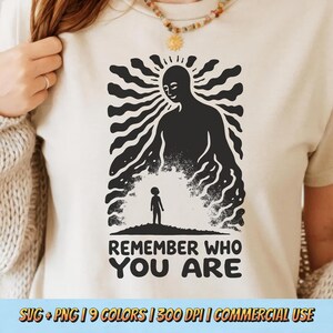 May include: Beige t-shirt featuring a black graphic of a large figure with a smaller figure below, with the text "REMEMBER WHO YOU ARE". The design includes sun-like rays and a grassy area. The shirt is a neutral color.