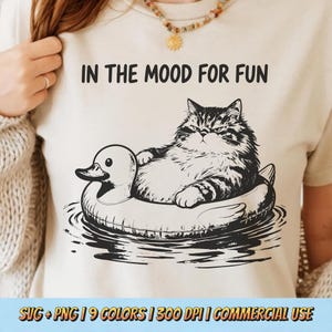 Könnte beinhalten: Cremefarbenes T-Shirt mit einer Schwarz-Weiß-Illustration einer Katze, die auf einem Enten-Schwimmring im Wasser liegt. Der Text "IN THE MOOD FOR FUN" steht über der Katze. Der untere Teil des Shirts hat den Text "SVG + PNG | 9 COLORS | 300 DPI | COMMERCIAL USE".