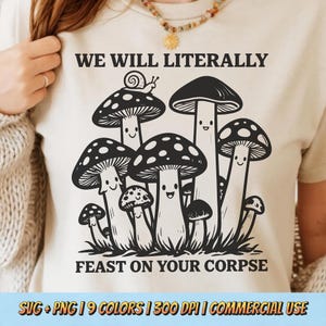 May include: Beige t-shirt with a black and white graphic of cartoon mushrooms with smiling faces. The text above the mushrooms reads "WE WILL LITERALLY" and below reads "FEAST ON YOUR CORPSE".