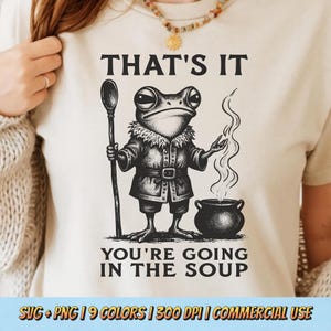 May include: Beige t-shirt featuring a whimsical illustration of a frog in a coat holding a spoon and gesturing towards a bubbling pot. The text "THAT'S IT YOU'RE GOING IN THE SOUP" is printed above and below the frog. The shirt also has the text "SVG + PNG | 9 COLORS | 300 DPI | COMMERCIAL USE".