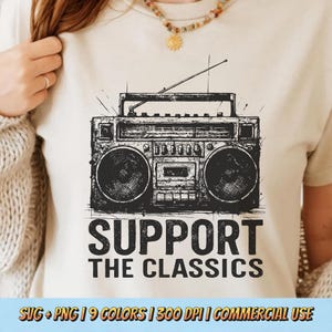 May include: Cream-colored t-shirt featuring a black and white illustration of a vintage boombox. The text "SUPPORT THE CLASSICS" is printed below the boombox. The shirt has a crew neck and short sleeves.