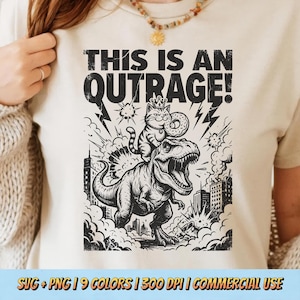 May include: Cream-colored t-shirt featuring a black and white graphic. The graphic depicts a cat wearing a crown and riding a Tyrannosaurus Rex, with the text "THIS IS AN OUTRAGE!". The design includes lightning bolts and city buildings.