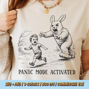 Puede incluir: Camiseta beige con una ilustración en blanco y negro de un niño huyendo de un conejo amenazante. El texto "PANIC MODE ACTIVATED" está impreso debajo. La camiseta tiene cuello redondo y mangas cortas.