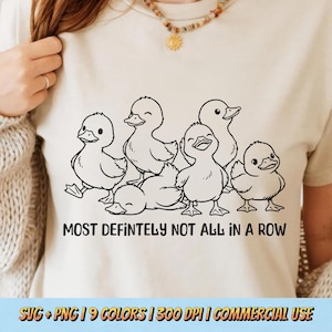 May include: A light beige t-shirt featuring a black and white illustration of six ducklings. The text "MOST DEFINITELY NOT ALL IN A ROW" is printed below the ducklings. The shirt is a casual style.