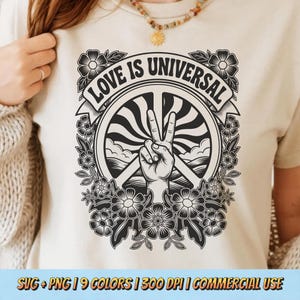 Puede incluir: Camiseta beige con un gráfico en blanco y negro. El gráfico incluye un gesto de la mano de la paz, un diseño circular con las palabras "LOVE IS UNIVERSAL" y elementos florales. La camiseta es de color neutro.