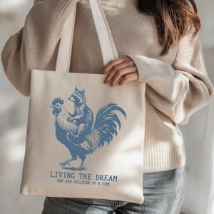 May include: Beige tote bag with a blue illustration of a raccoon riding a rooster. The text "LIVING THE DREAM ONE BAD DECISION AT A TIME" is printed below the image. The bag has two long handles.
