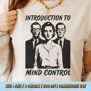 May include: Beige t-shirt with a black and white graphic of three people and the text "INTRODUCTION TO MIND CONTROL". The graphic features a woman and two men in suits. The shirt is a neutral color.
