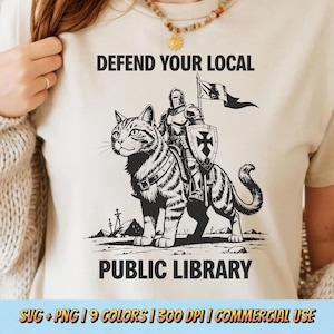 May include: Beige t-shirt with a black and white illustration of a knight riding a large, striped cat. The text "DEFEND YOUR LOCAL PUBLIC LIBRARY" is above and below the image. The bottom of the shirt has text that reads "SVG + PNG | 9 COLORS | 300 DPI | COMMERCIAL USE".