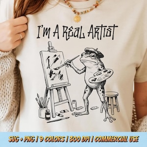 May include: Beige t-shirt with the text "I'M A REAL ARTIST" in black. The shirt features a black and white illustration of a frog wearing a beret, holding a paintbrush and palette, painting on a canvas. Additional art supplies are also shown.