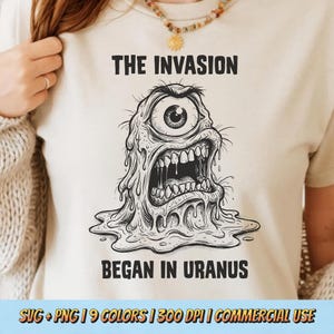 May include: A beige t-shirt featuring a black ink-style illustration of a one-eyed, melting alien creature. Above the creature, the text reads THE INVASION, and below it, the text reads BEGAN IN URANUS. The design is centered on the chest.