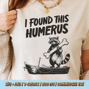 May include: Beige t-shirt with the text "I FOUND THIS HUMERUS" in black. A cartoon raccoon holding a bone stands in a small boat. The design is in black and white.