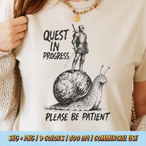 Puede incluir: Camiseta color crema con una ilustración en blanco y negro de un caballero sobre un caracol. El texto "QUEST IN PROGRESS" y "PLEASE BE PATIENT" está presente. La camiseta es de color neutro claro.