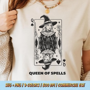 Op de afbeelding: Crèmekleurig T-shirt met een zwart-wit illustratie van een schoppenkoningin kaart. De kaart toont een vrouw met een heksenhoed die een toverstaf en een bloem vasthoudt. De tekst "QUEEN OF SPELLS" staat eronder.