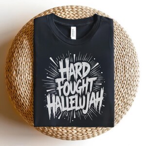 May include: Black t-shirt with the words "HARD FOUGHT HALLELUJAH" in a white, stylized font. The text is surrounded by radiating lines, creating a burst effect. The shirt is displayed on a round, woven surface.