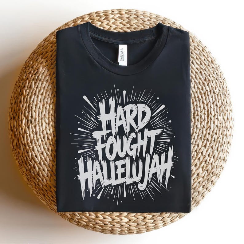 May include: Black t-shirt with the words "HARD FOUGHT HALLELUJAH" in a white, stylized font. The text is surrounded by radiating lines, creating a burst effect. The shirt is displayed on a round, woven surface.