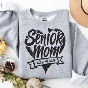 May include: A light gray sweatshirt with the words "Senior Mom" in a black heart-shaped design. Below the heart is a banner that reads "class of 2026". The sweatshirt has white cuffs and a white interior.