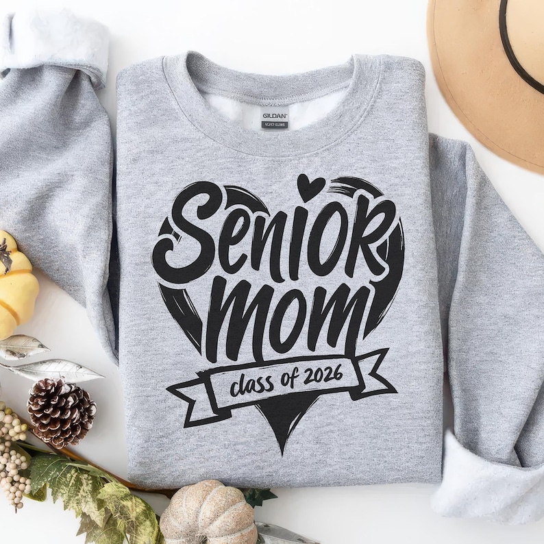 May include: A light gray sweatshirt with the words "Senior Mom" in a black heart-shaped design. Below the heart is a banner that reads "class of 2026". The sweatshirt has white cuffs and a white interior.