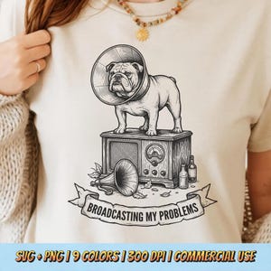 Peut inclure: T-shirt crème avec une illustration détaillée d'un bouledogue portant une collerette, debout sur une radio vintage. Une bannière en dessous indique "BROADCASTING MY PROBLEMS". Le design comprend un mégaphone et des bouteilles. Le texte inférieur indique "SVG + PNG | 9 COLORS | 300 DPI | COMMERCIAL USE."
