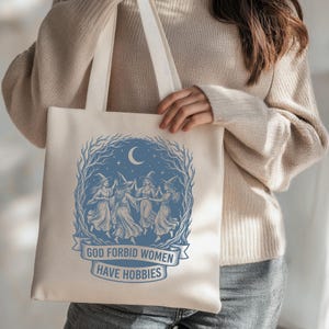 May include: Beige tote bag with a blue illustration of witches dancing under a crescent moon and stars. The text "GOD FORBID WOMEN HAVE HOBBIES" is printed on a banner below the illustration. The bag has long, cream-colored handles.