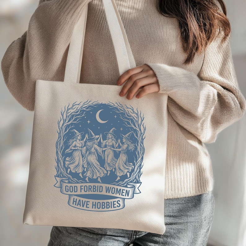 May include: Beige tote bag with a blue illustration of witches dancing under a crescent moon and stars. The text "GOD FORBID WOMEN HAVE HOBBIES" is printed on a banner below the illustration. The bag has long, cream-colored handles.