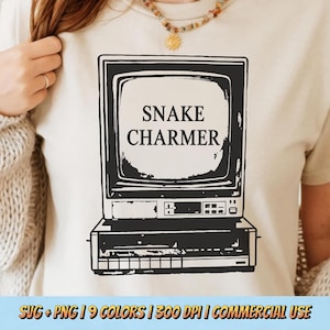 May include: A cream-colored t-shirt featuring a black graphic of a vintage television set with the text SNAKE CHARMER displayed on the screen. The design includes detailed buttons and a base unit, presented in a high-contrast, screen-printed style.