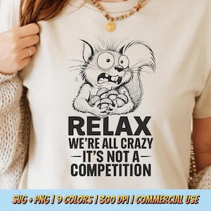 Può includere: Una maglietta color crema con un disegno in bianco e nero di uno scoiattolo dei cartoni animati che tiene delle ghiande, con un'espressione di panico. Sotto, il testo recita: "RELAX WE'RE ALL CRAZY - IT'S NOT A - COMPETITION".